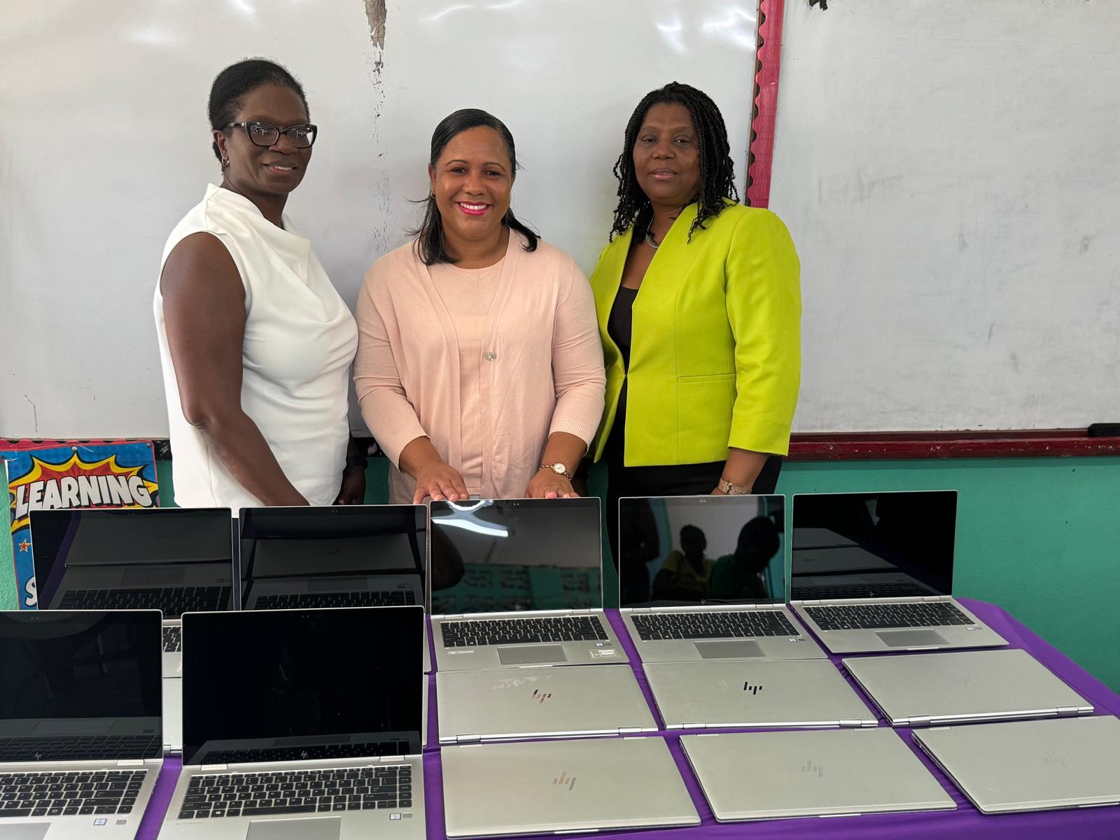 Three women stand behind a table with several laptops, smiling. The scene highlights a technology donation or educational initiative in a classroom.