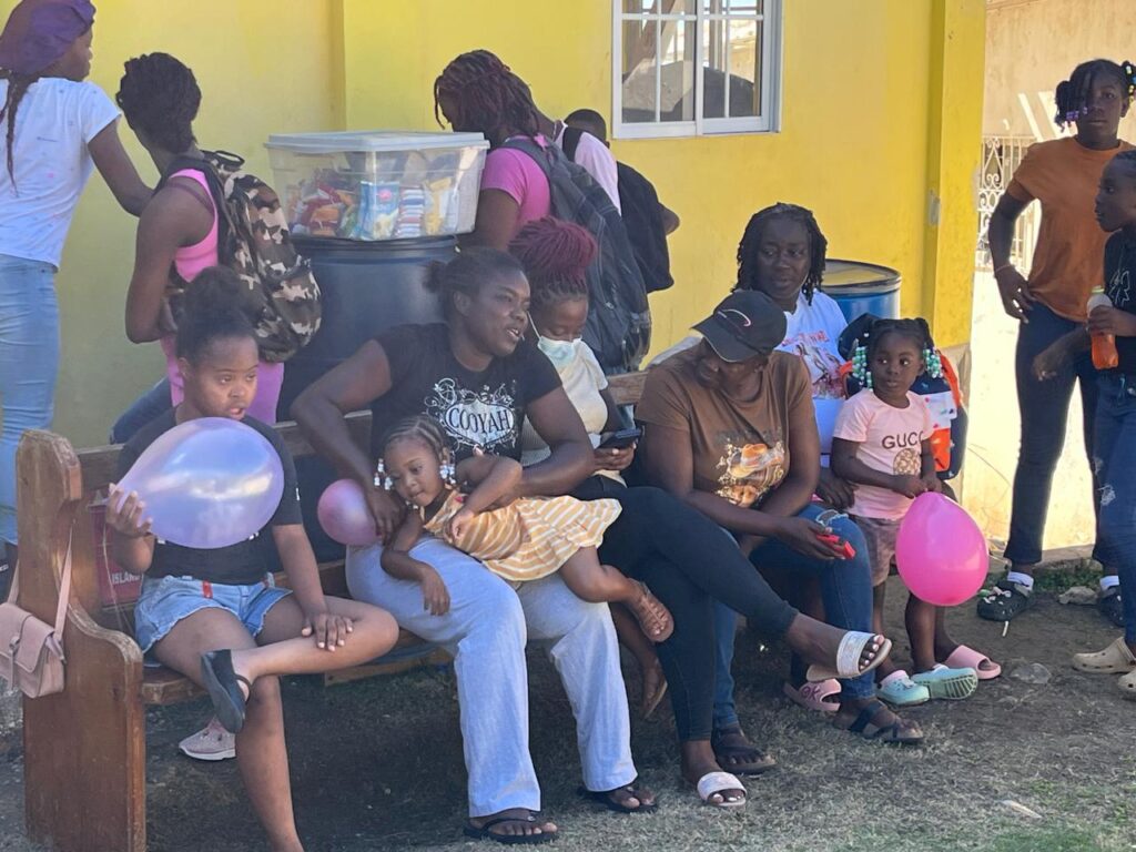 A group of women and children gather outside a yellow building, some holding balloons. They appear engaged in conversation and celebration.