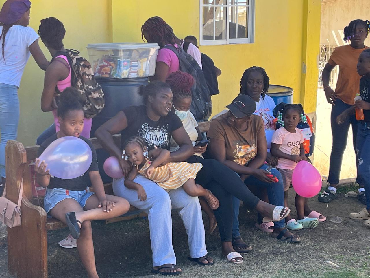 A group of women and children gather outside a yellow building, some holding balloons. They appear engaged in conversation and celebration.