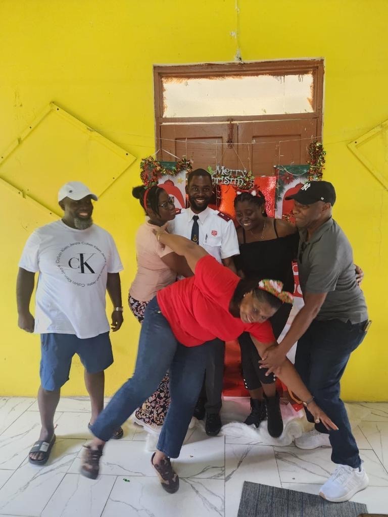 A group of five people joyfully posing in front of a vibrant yellow wall, celebrating a holiday event with laughter and playful interactions.