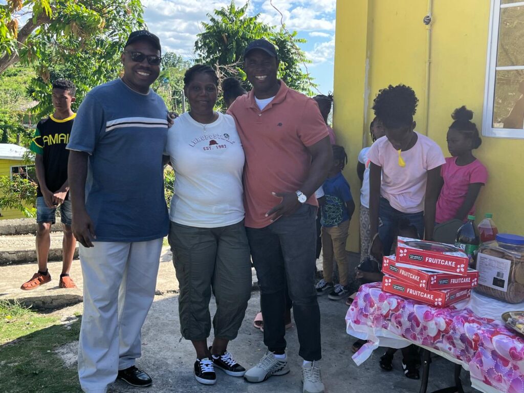 Three adults smile for a photo in front of a yellow building, surrounded by children and a table with fruitcake and refreshments, celebrating a community event.