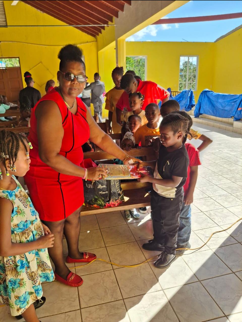 A woman in a red dress distributes gifts to excited children in a bright yellow room, celebrating a special event together.