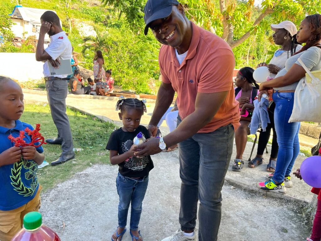A smiling man hands a toy to a young girl wearing a black shirt, while another child holds red toys. Friends and family gather in a green outdoor setting.