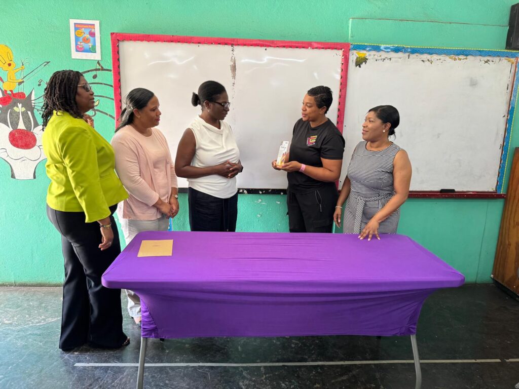 Five women engage in a discussion around a purple table in a colorful classroom. One woman holds a product while others listen attentively.