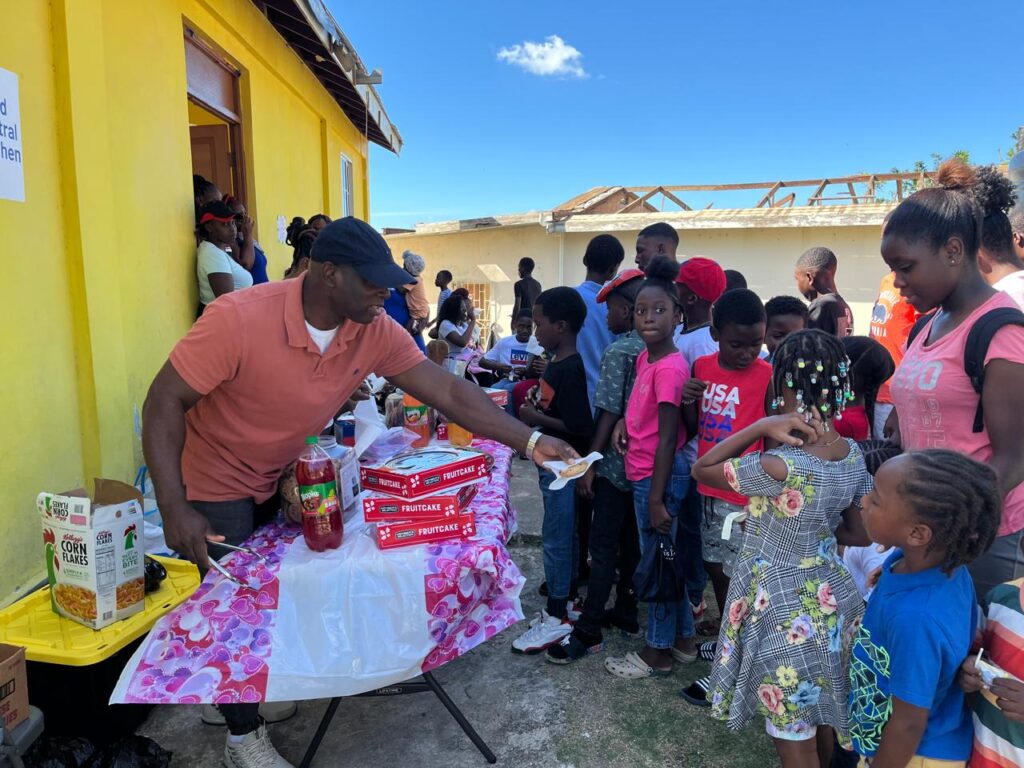 A man distributes food at a community event outside a yellow building, while a line of children eagerly waits for their turn.