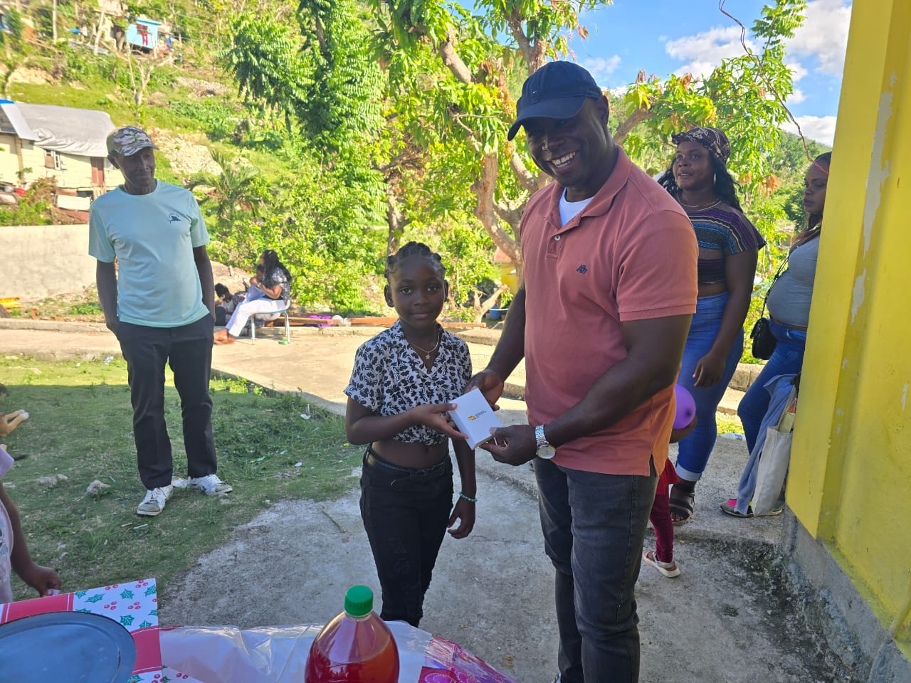 A smiling man hands a gift to a young girl in a casual outdoor setting. Family and friends gather nearby, celebrating a special occasion.