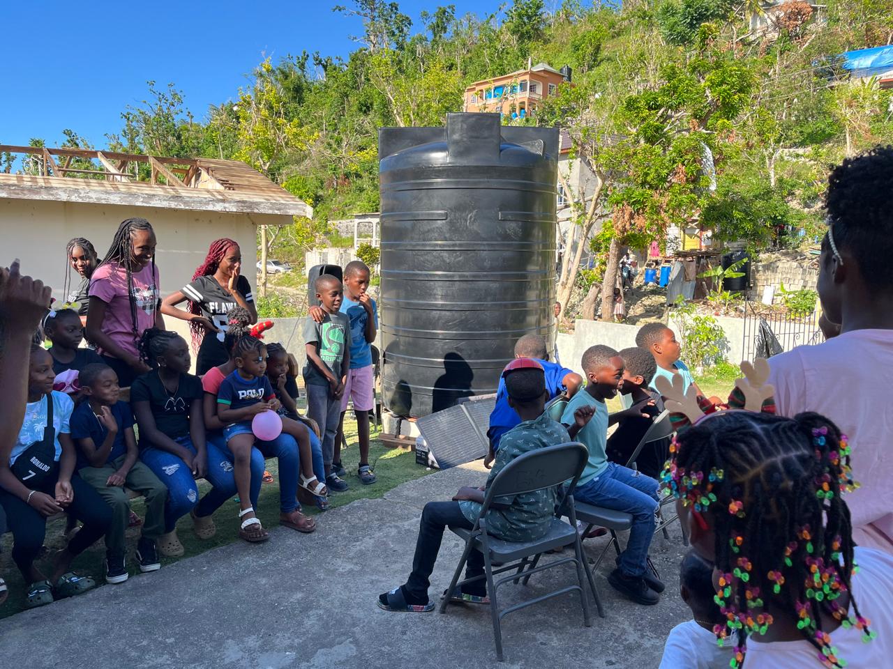 Children gather in an outdoor space for a fun event, sitting on chairs near a large water tank. They appear engaged and joyful, creating a lively atmosphere.