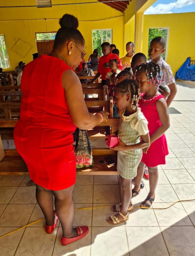 A woman in a red dress hands a small gift to a child in a light dress, surrounded by other children in a vibrant, sunny room.