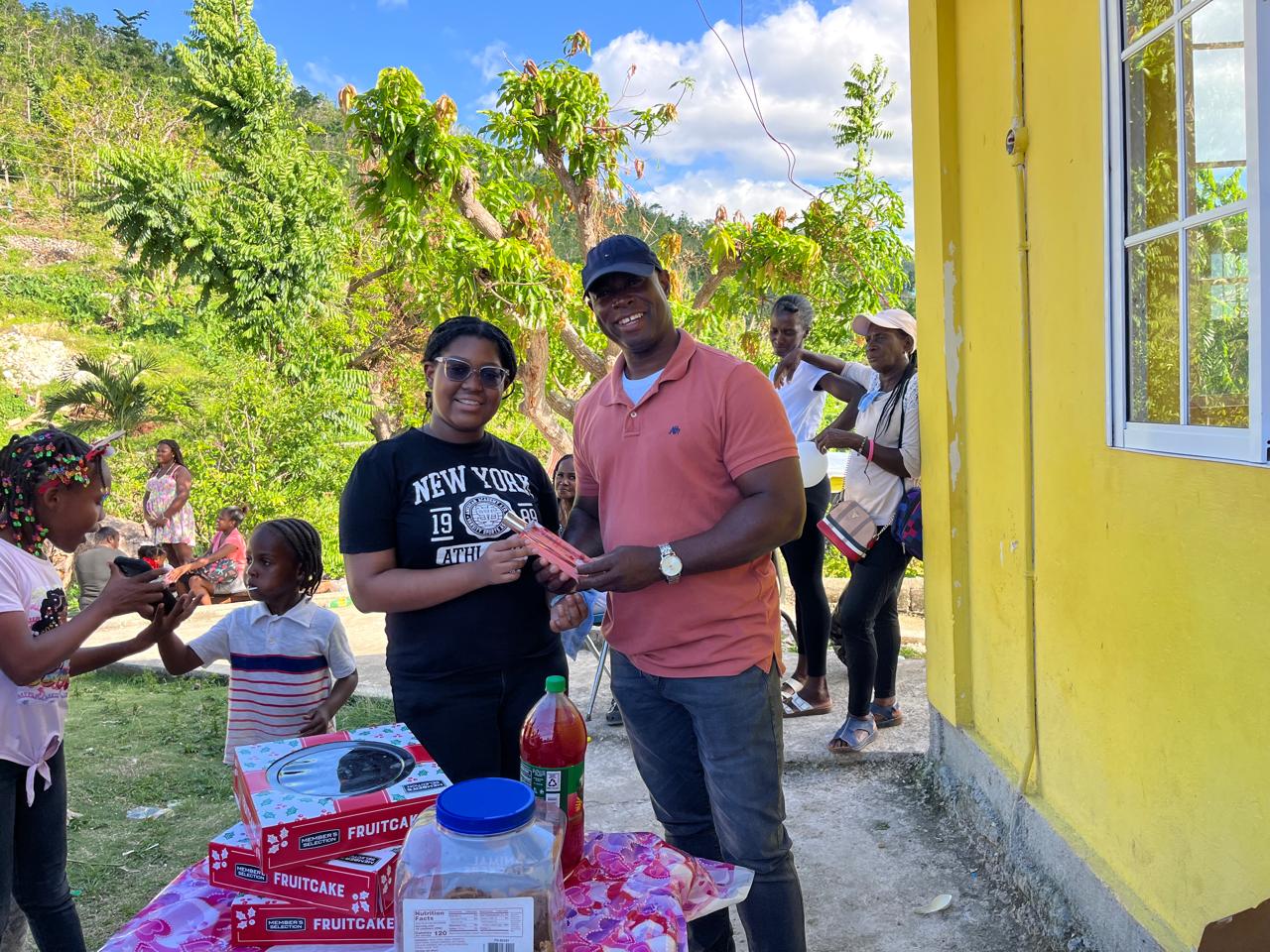 A man and a woman exchange items in front of a table with fruitcakes at a vibrant outdoor gathering, surrounded by smiling children and adults.