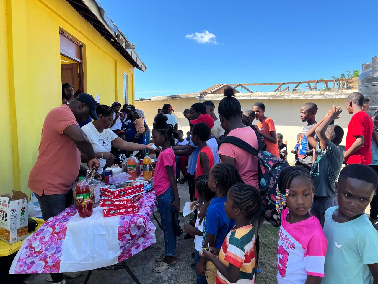 Children gather outside a yellow building for a community event, waiting in line as adults serve food and drinks from a table with snacks.