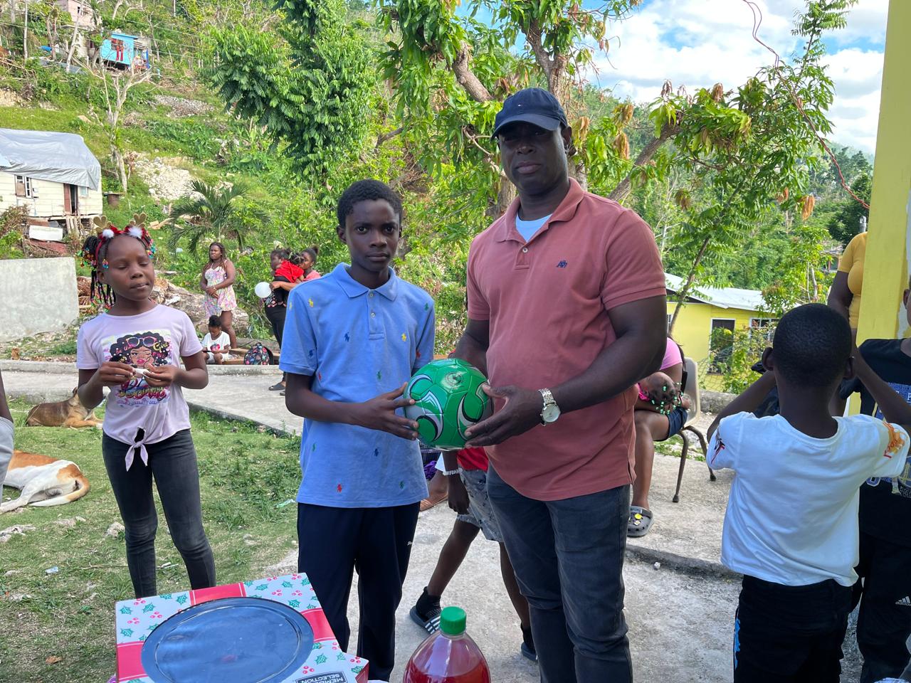 A child receives a green soccer ball from a man in a pink polo shirt at a community gathering, with other children and activities visible in the background.