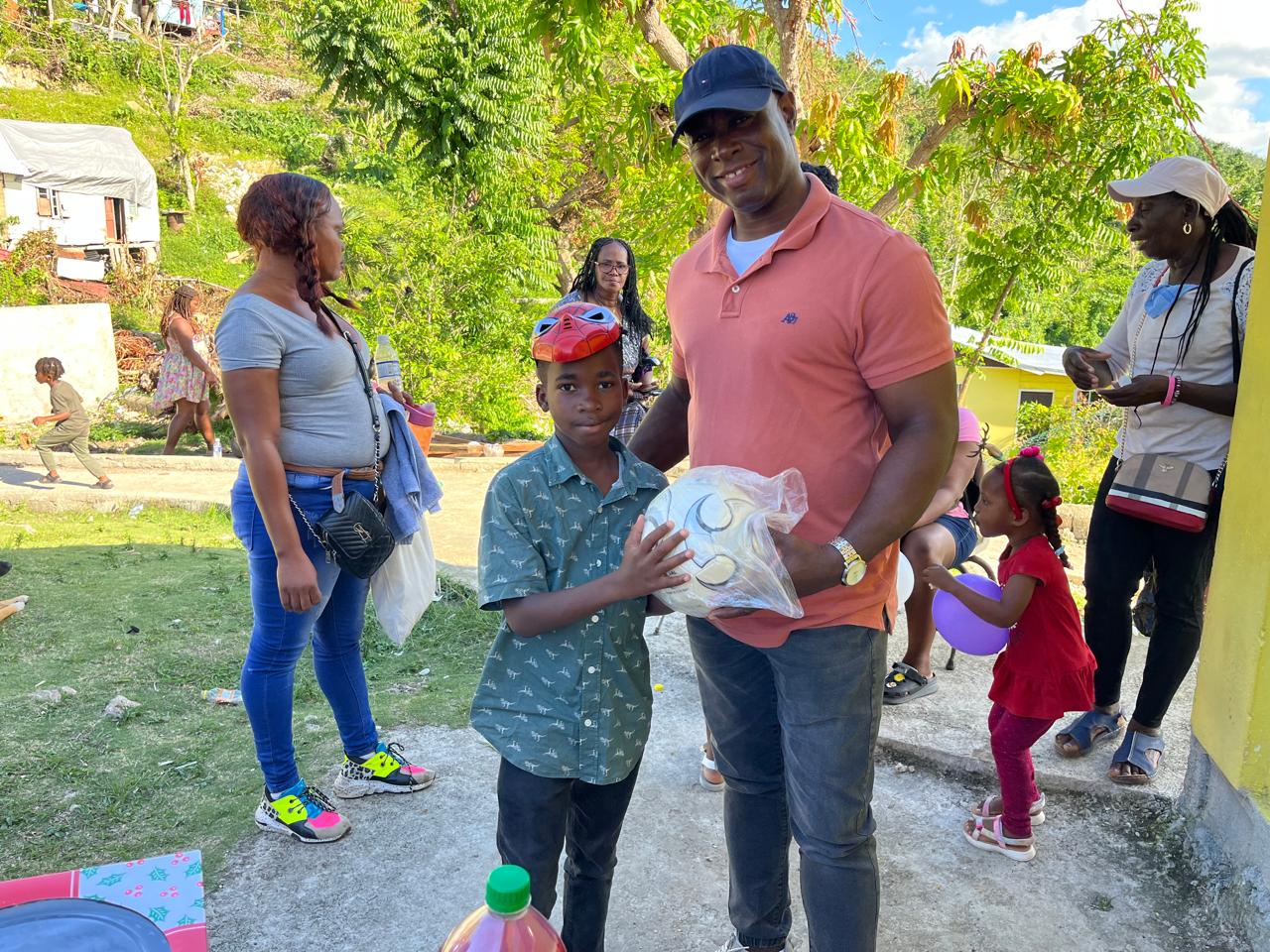 A man in an orange shirt presents a wrapped gift to a boy wearing a red cap. The scene features children and adults enjoying a festive gathering outdoors.