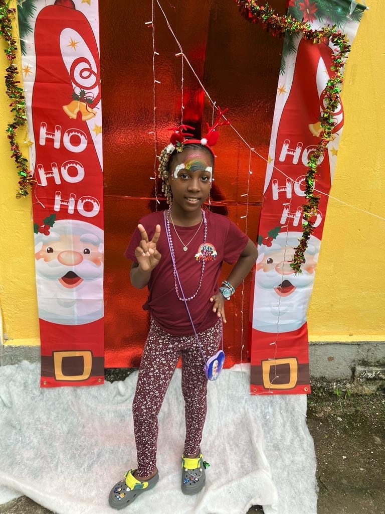Girl dressed in festive attire with reindeer antlers and face paint, posing cheerfully in front of a decorated holiday backdrop.