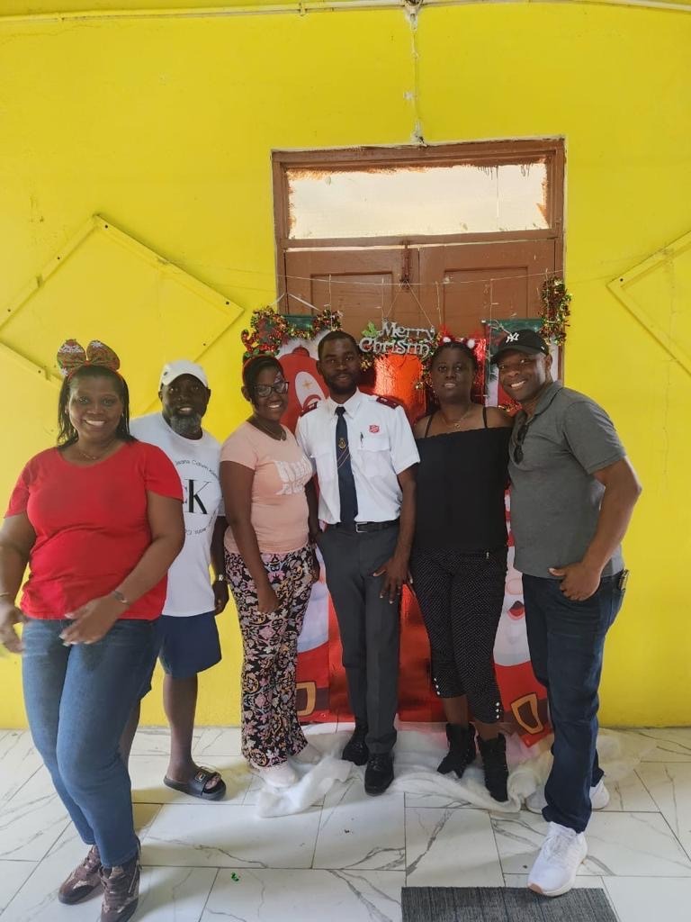 Group of six people posing in front of a festive, yellow wall decorated with a "Merry Christmas" sign, celebrating the holiday together.