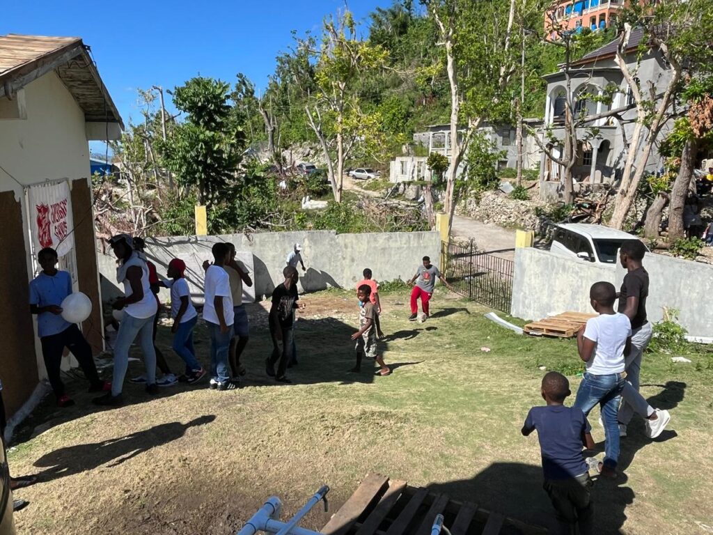 Children play in a yard, some holding balloons, with a backdrop of damaged trees and buildings. The scene reflects community resilience and joy.