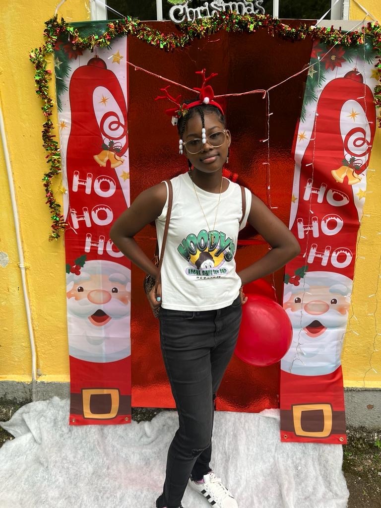 A girl with styled red hair and glasses poses in front of a festive Christmas backdrop featuring Santa decorations and a red balloon.