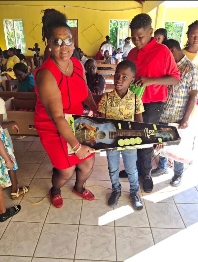 A woman in a red dress hands a child a new guitar in a community gathering. Others watch in the background, celebrating the occasion.