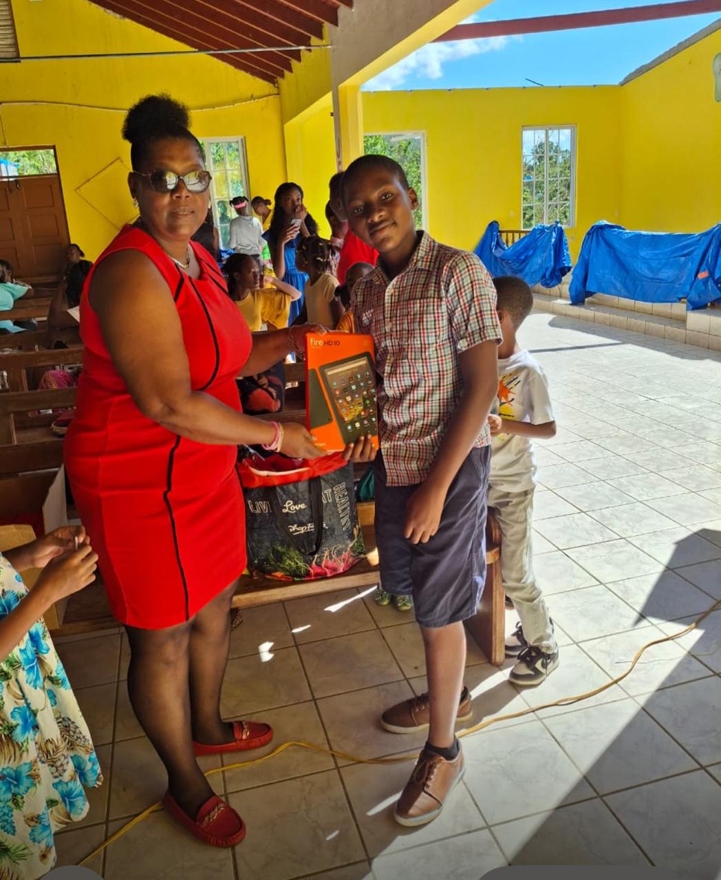 A woman in a red dress hands a boy a Fire HD tablet in a bright communal space, surrounded by children eagerly watching the gift exchange.
