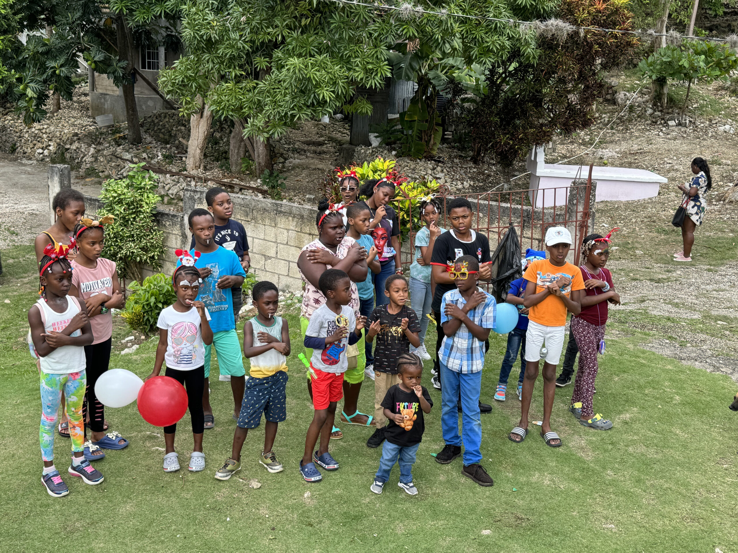 A group of children gathered outdoors, some holding balloons, celebrating an event with festive attire and accessories.