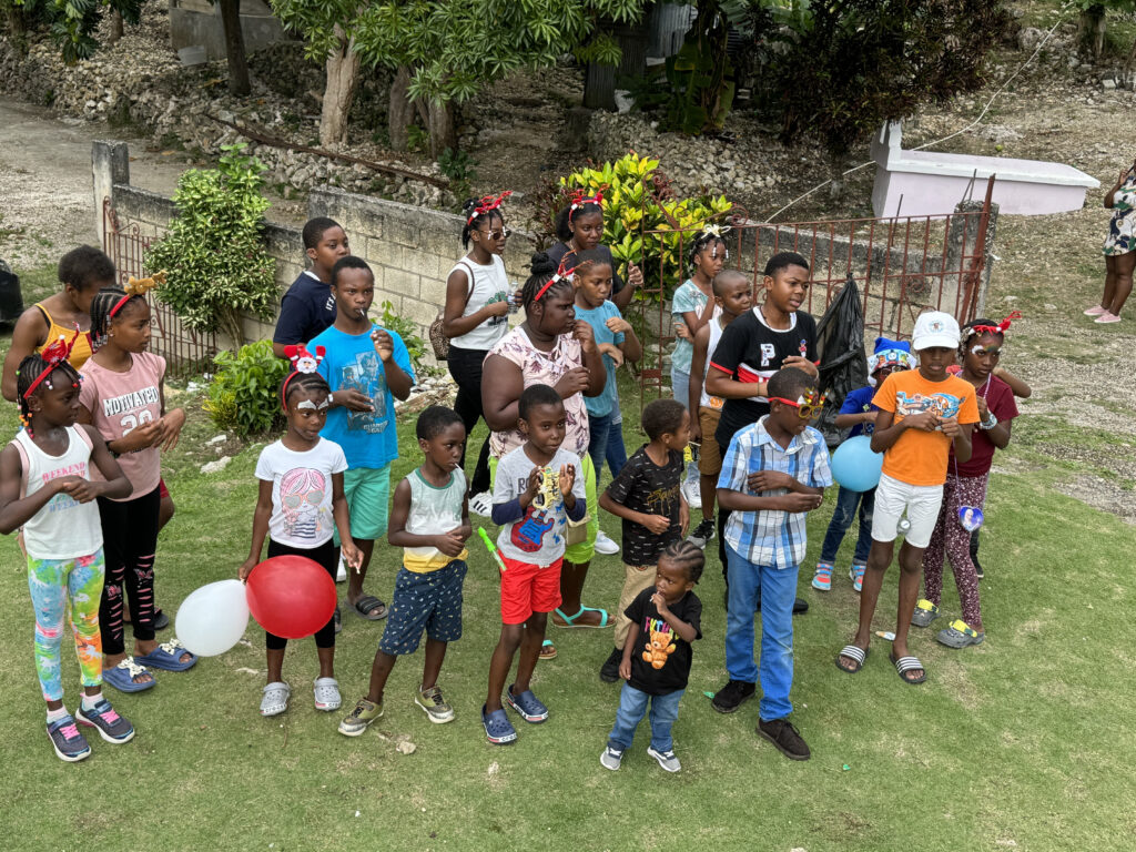A diverse group of children gathers outdoors, some holding balloons and wearing festive accessories, celebrating an event with joy and excitement.