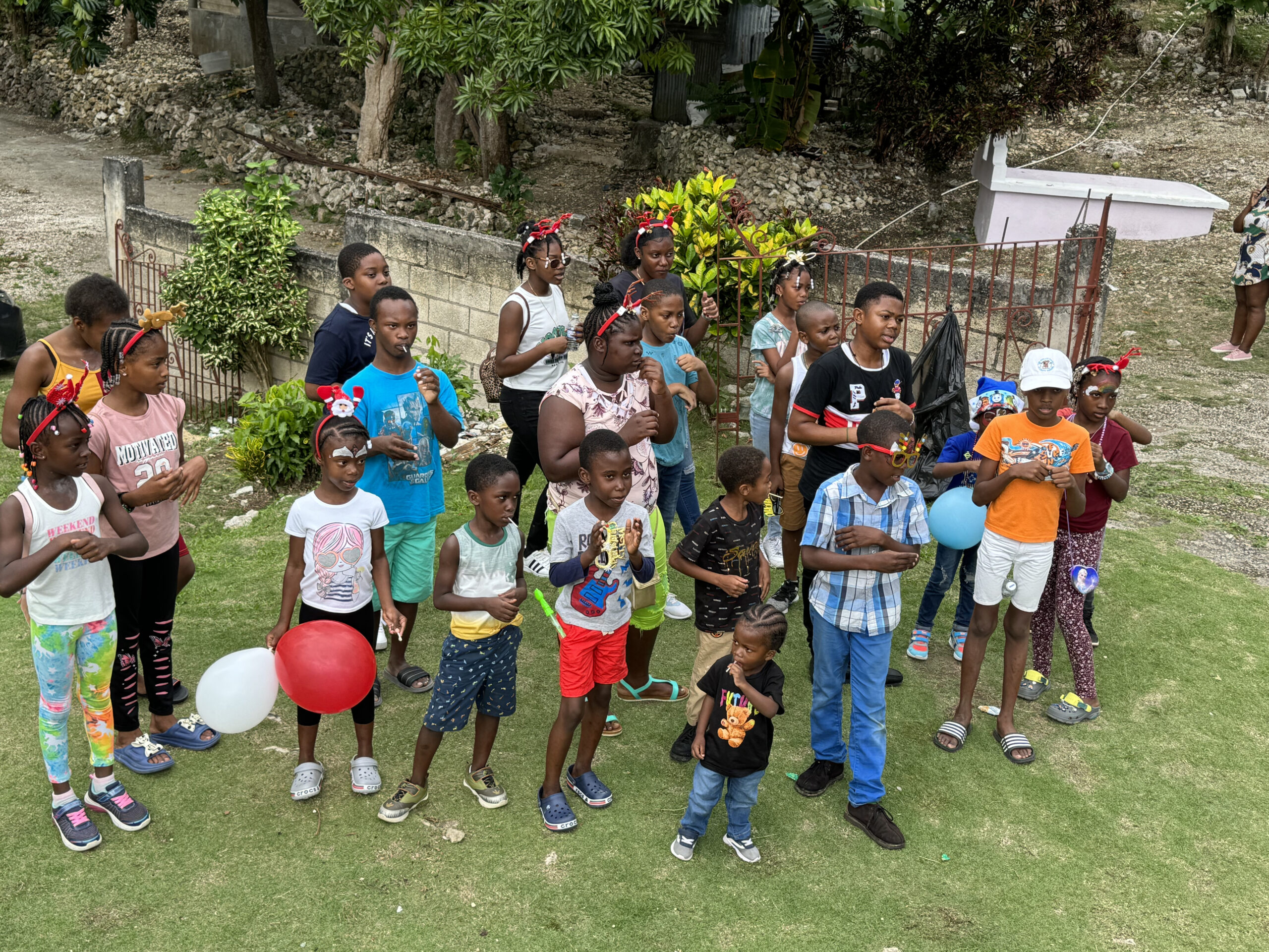 A diverse group of children gathers outdoors, some holding balloons and wearing festive accessories, celebrating an event with joy and excitement.