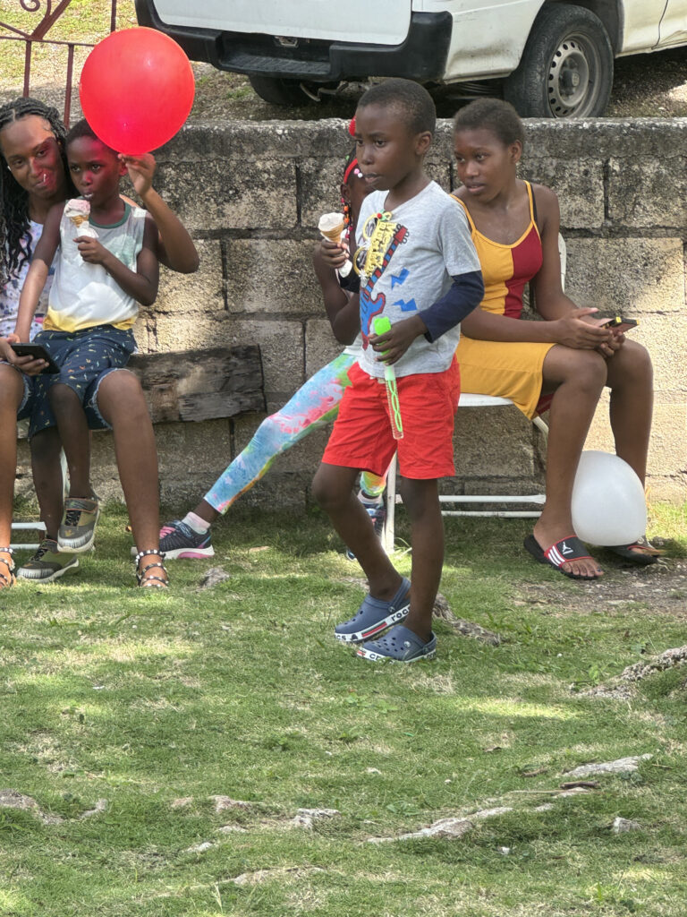 Children enjoy a sunny outdoor gathering, holding ice cream cones and balloons. A boy in red shorts stands apart, looking curious amidst joyful friends.