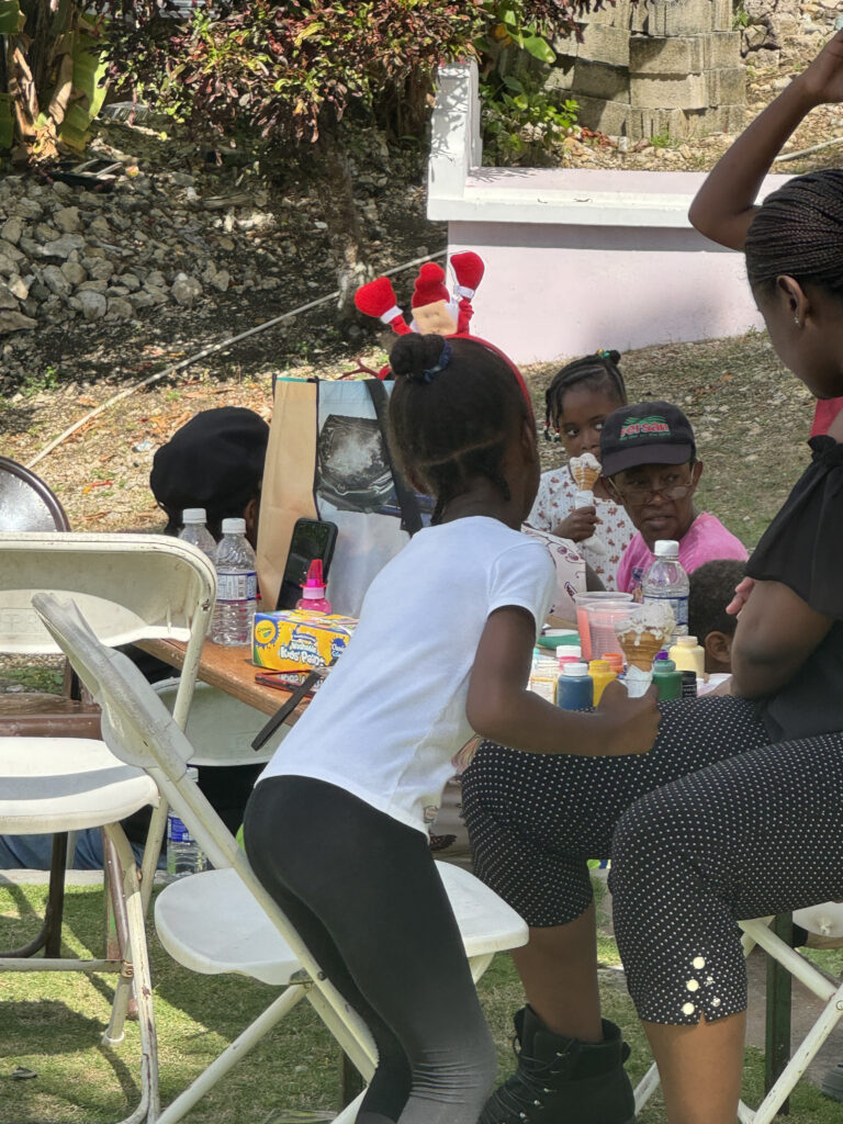 Children enjoying an outdoor activity, with a girl wearing a white shirt and black leggings painting at a table surrounded by adults and colorful materials.