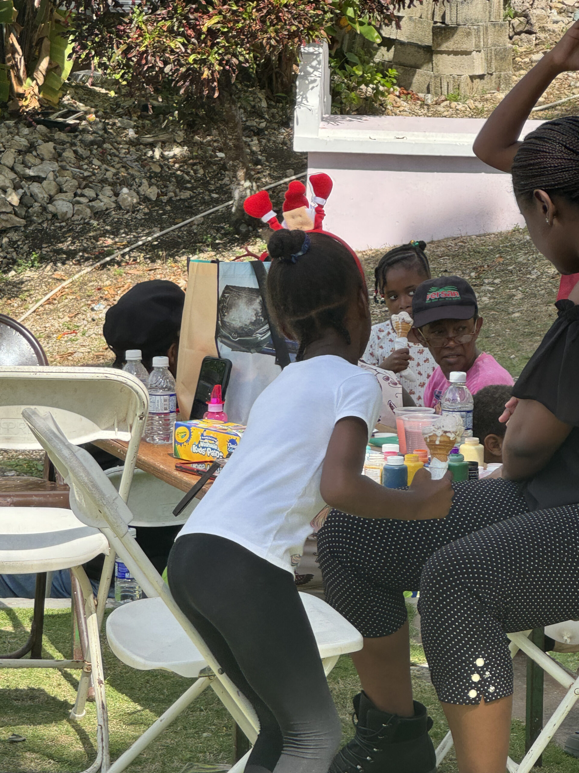 Children enjoying an outdoor activity, with a girl wearing a white shirt and black leggings painting at a table surrounded by adults and colorful materials.