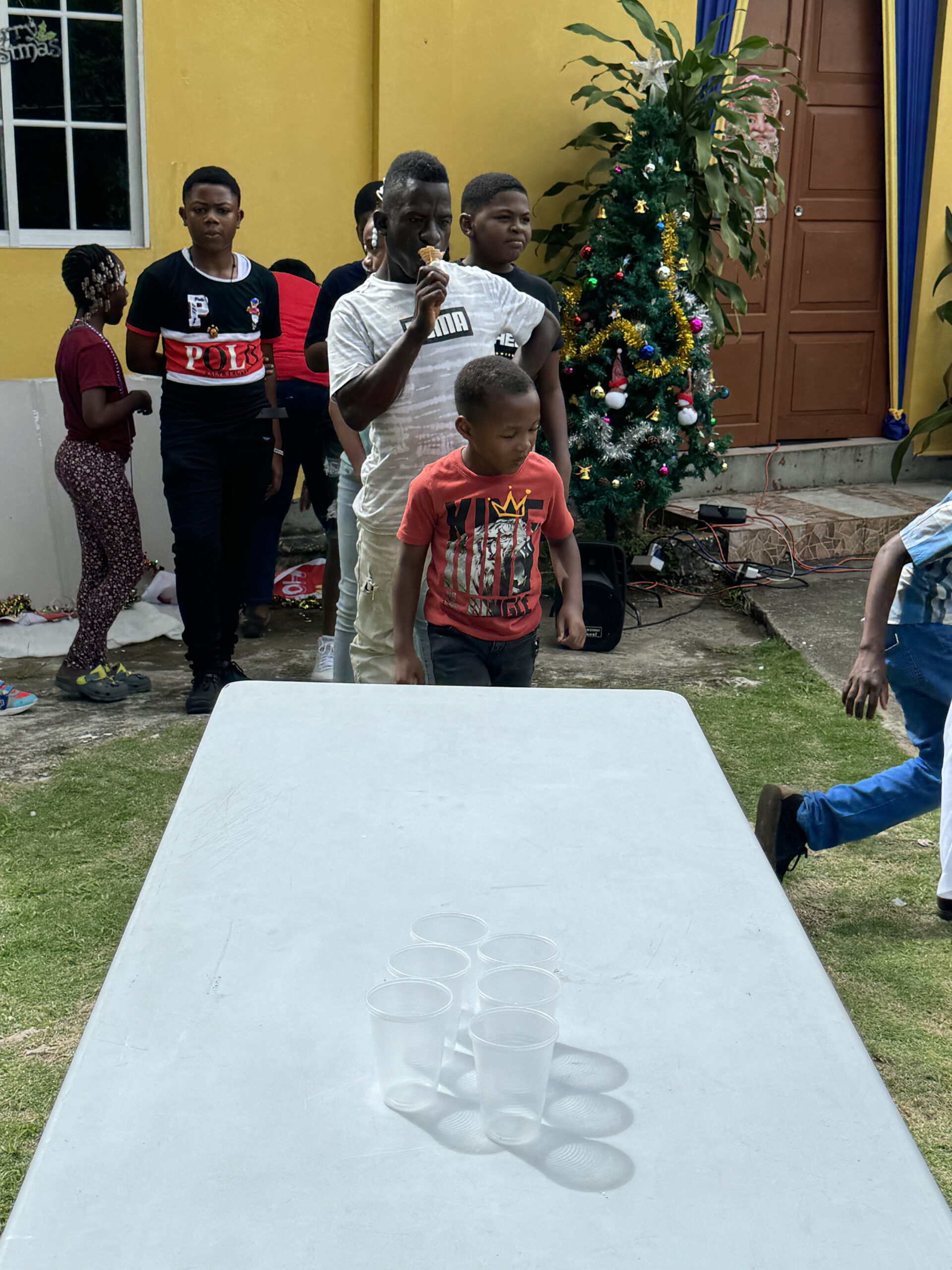 Children gather around a table with plastic cups during a festive outdoor gathering, with a decorated Christmas tree in the background.