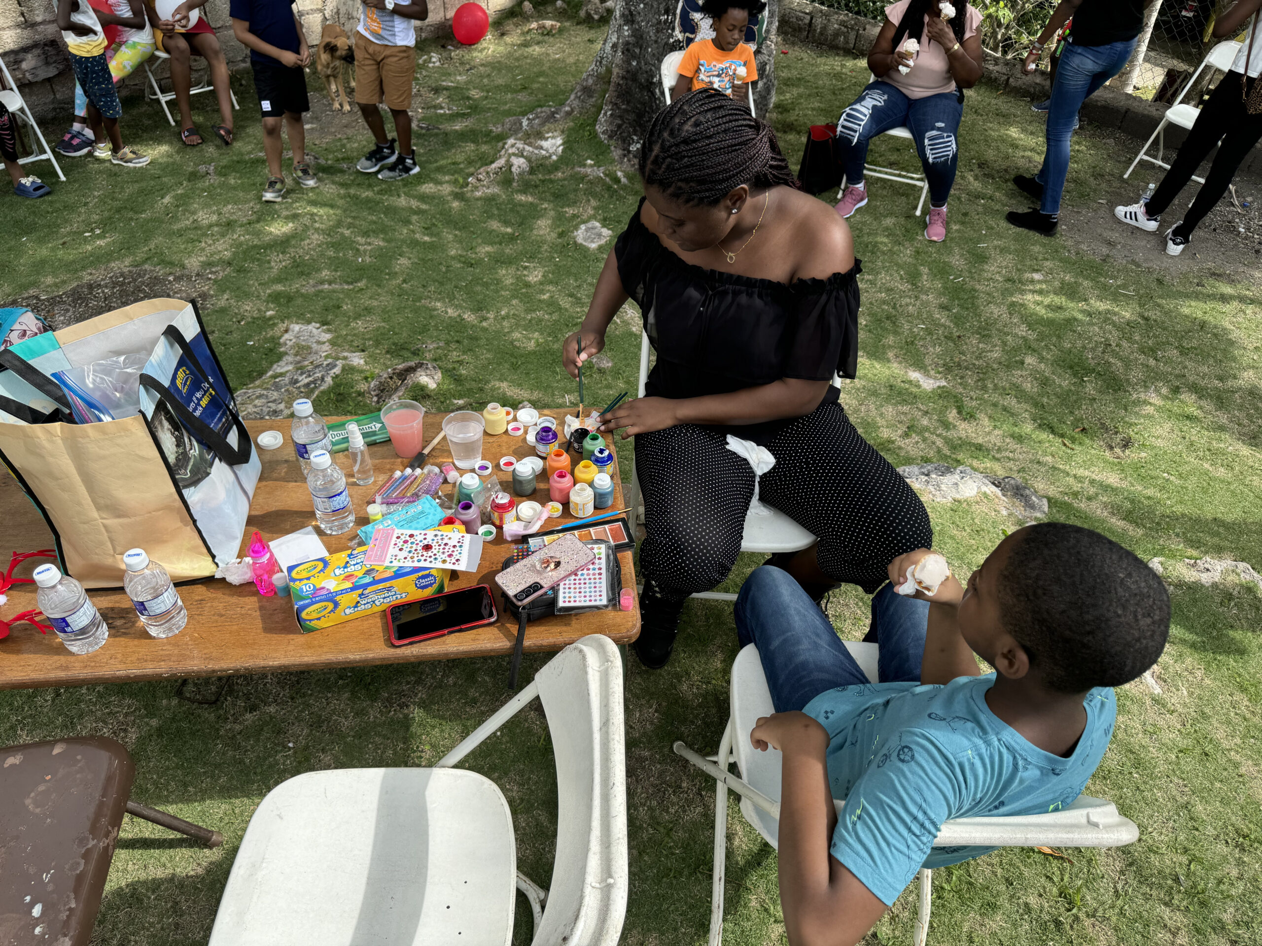 A woman paints a child's face at an outdoor event, surrounded by colorful art supplies. Nearby, children watch and enjoy refreshments.