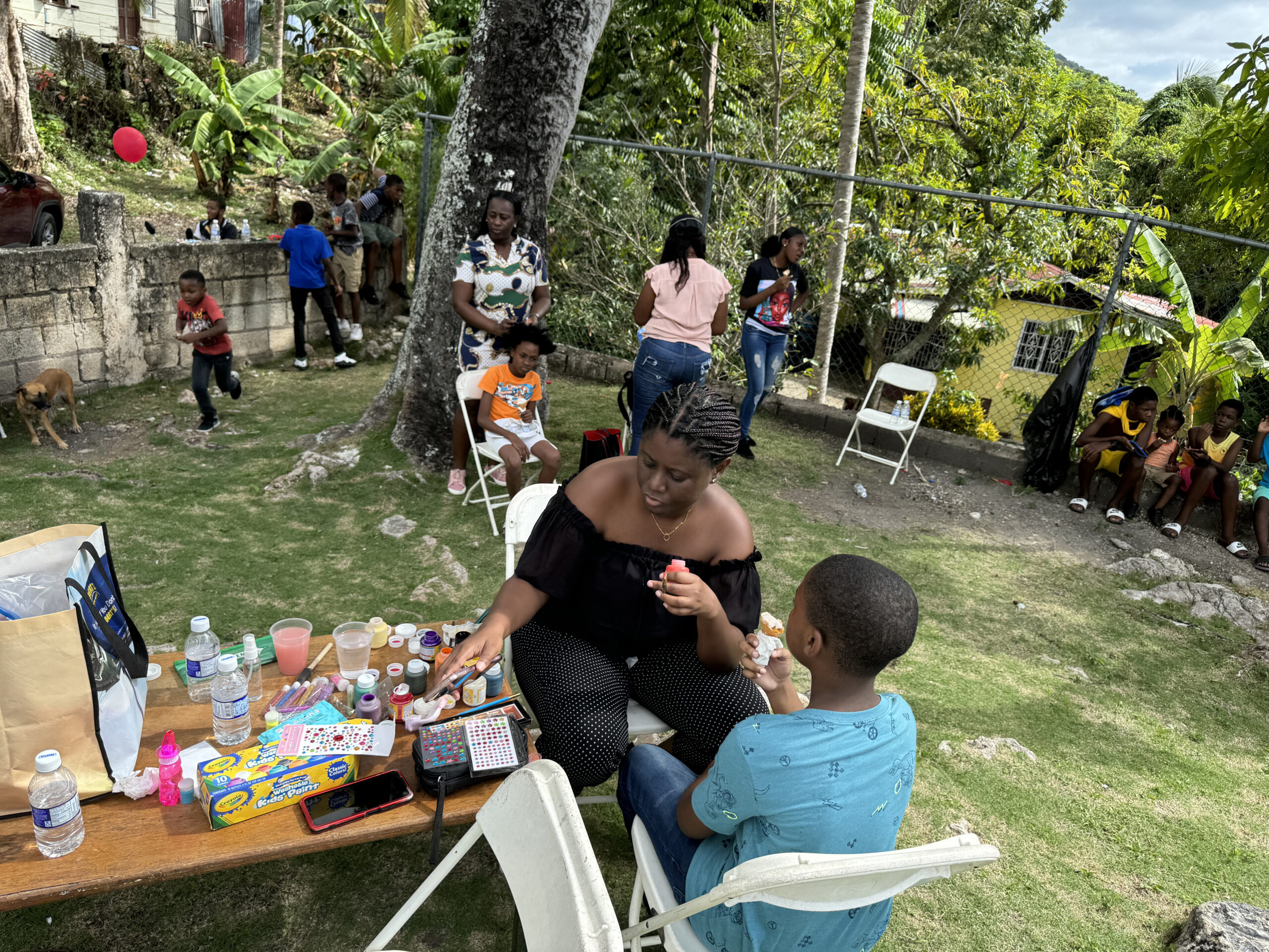 A woman applies face paint to a child at a festive outdoor gathering, surrounded by children playing and adults socializing in a lively, green setting.