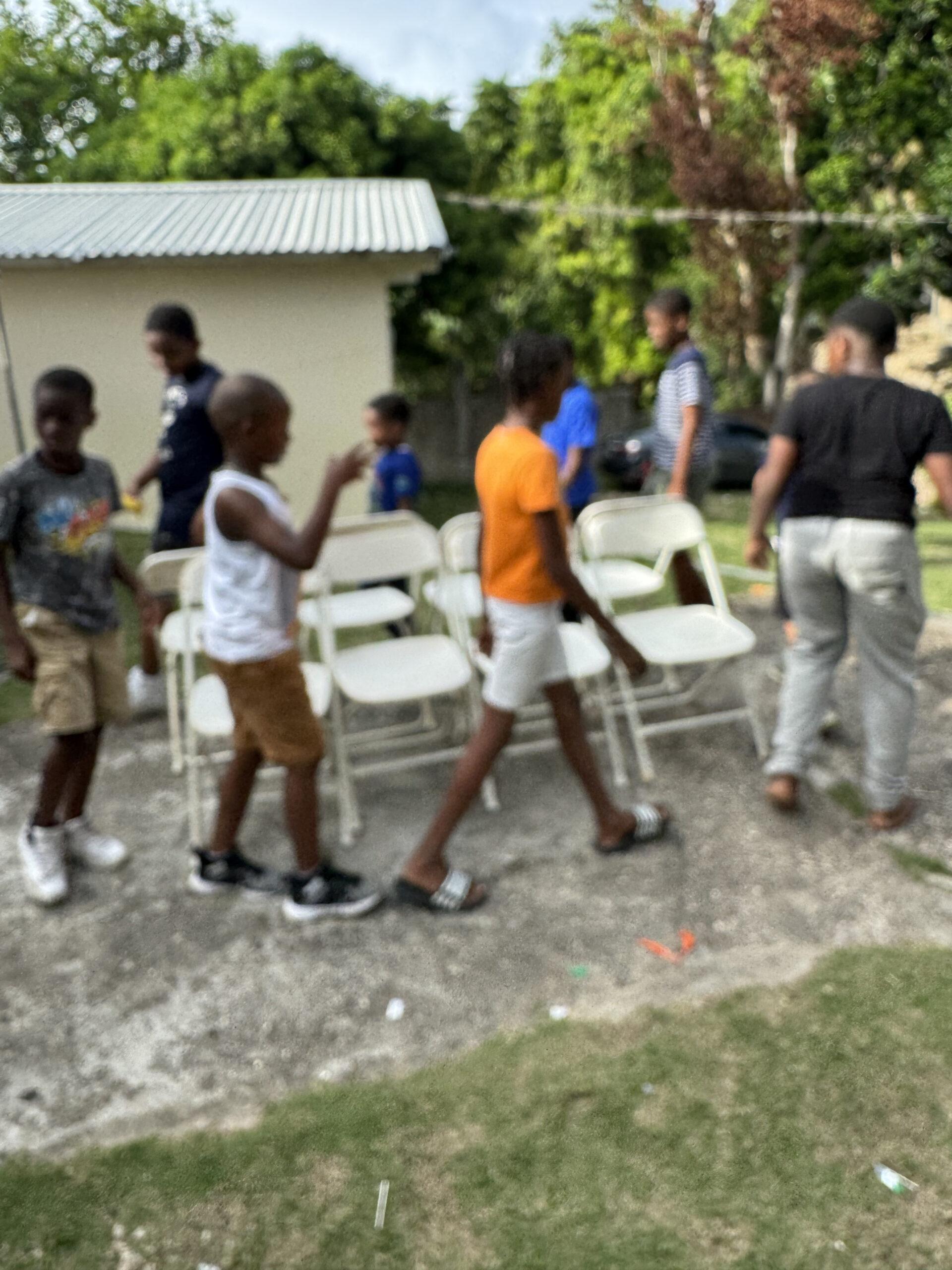 Children are actively moving folding chairs in a yard, preparing for an event, with a house and greenery in the background.