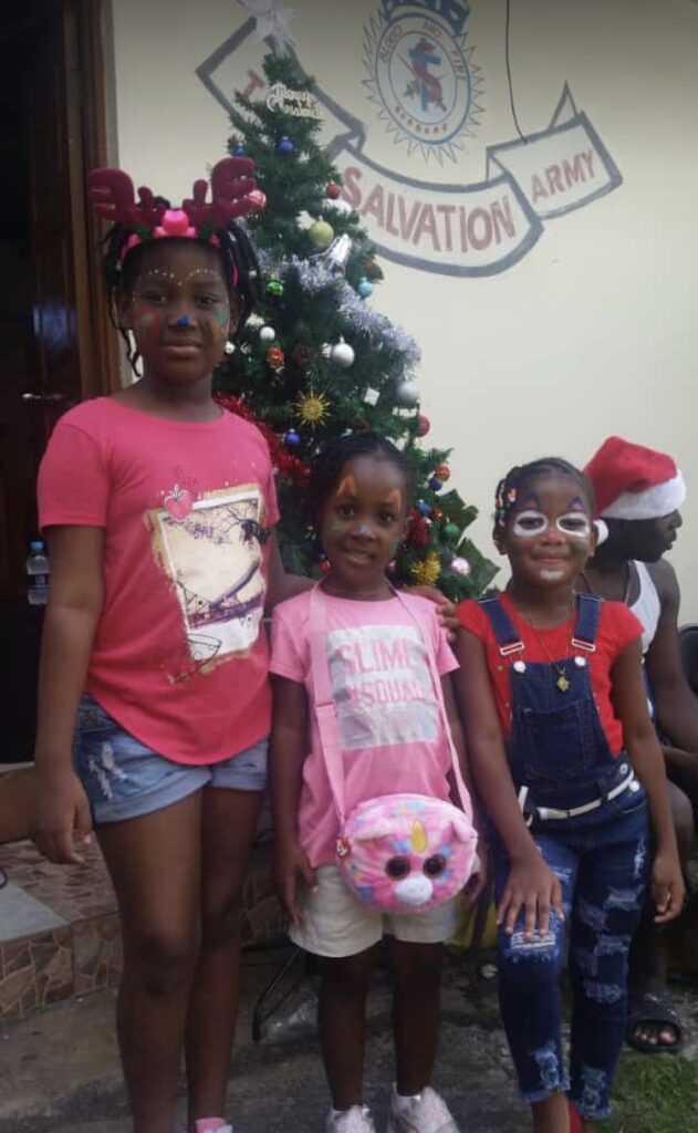 Three children celebrate near a decorated Christmas tree at a Salvation Army event. They wear festive clothing and face paint, smiling joyfully.