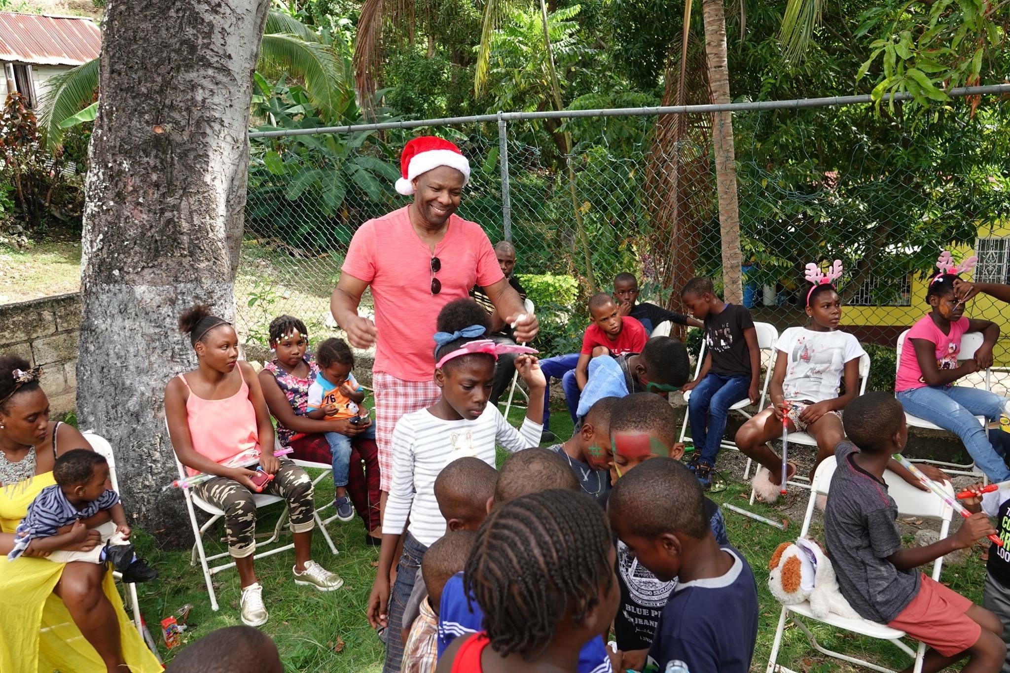 A man in a Santa hat engages with children during a festive gathering in a lush outdoor setting, surrounded by joyful participants of all ages.