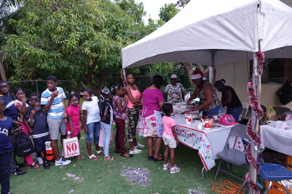 Children wait in line at a festive outdoor event, eyeing food being prepared under a decorated tent, creating a community gathering atmosphere.