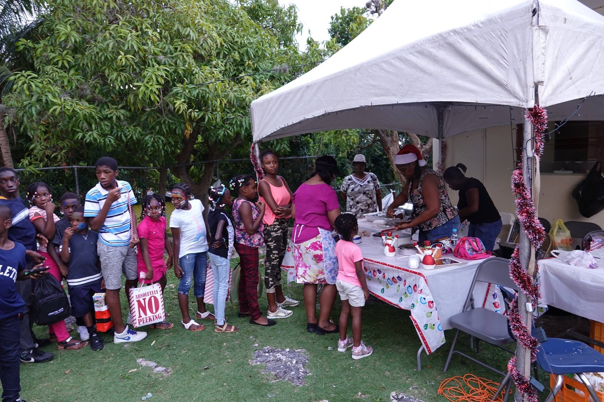 Children wait in line at a festive outdoor event, eyeing food being prepared under a decorated tent, creating a community gathering atmosphere.