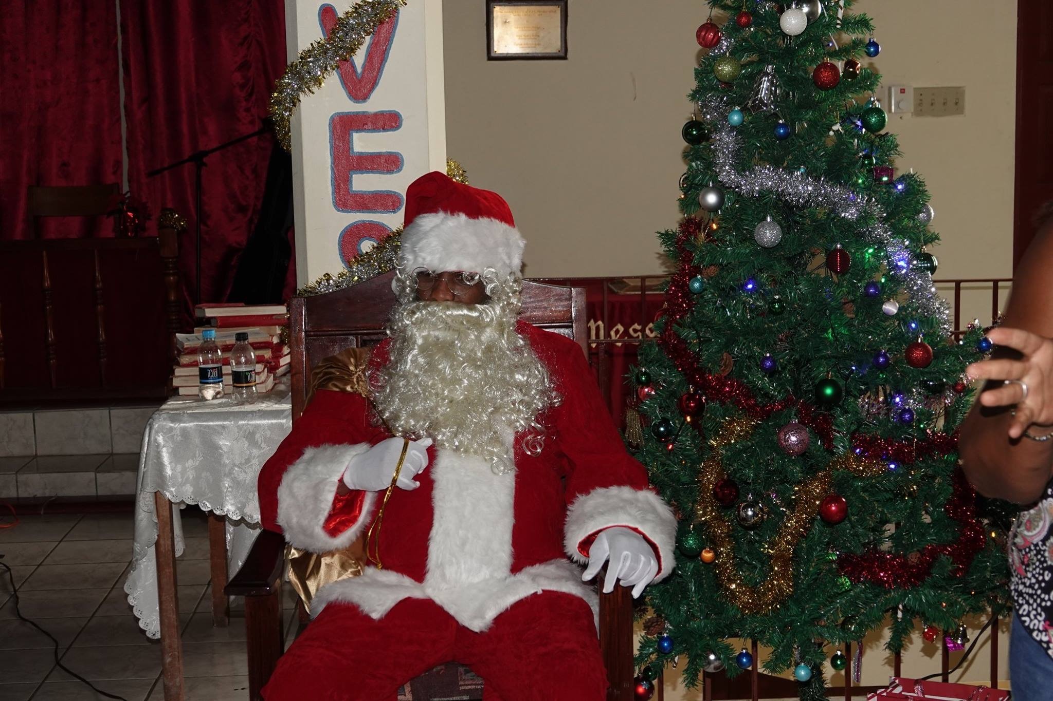 Santa Claus sits in a wooden chair beside a decorated Christmas tree, surrounded by festive ornaments and a warm holiday atmosphere.