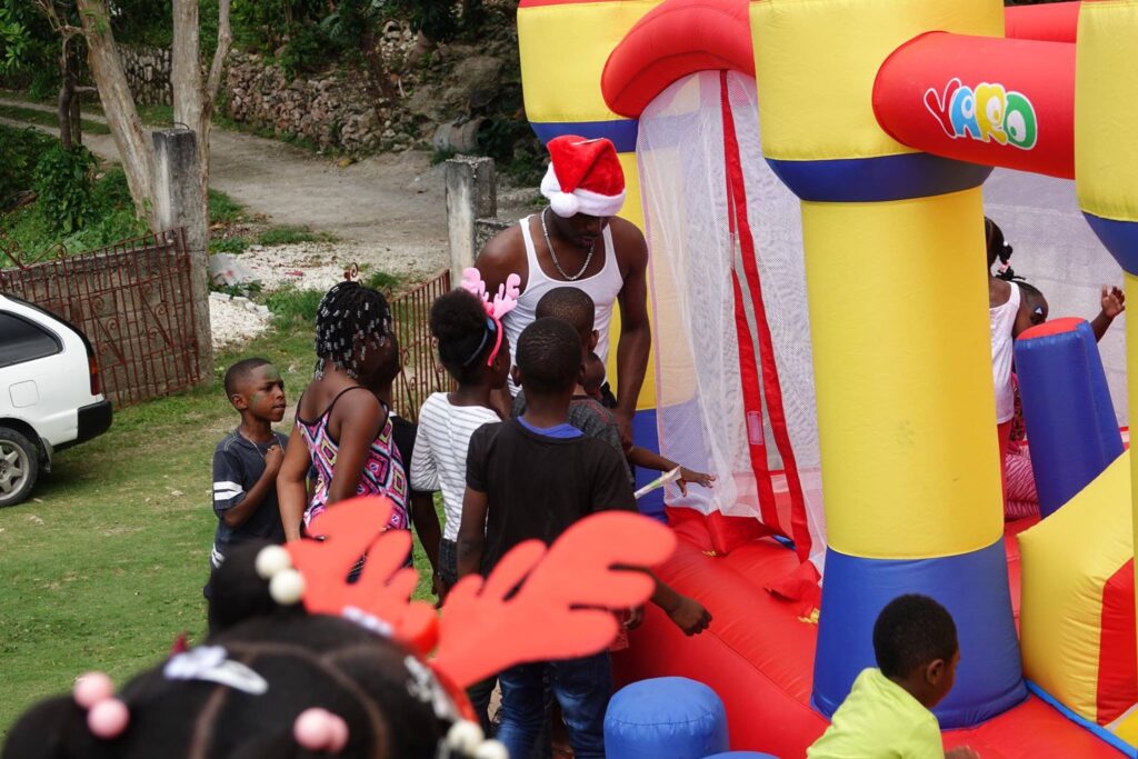 Children gather around a bounce house, excitement visible as a man in a Santa hat prepares to welcome them inside, fostering a festive atmosphere.