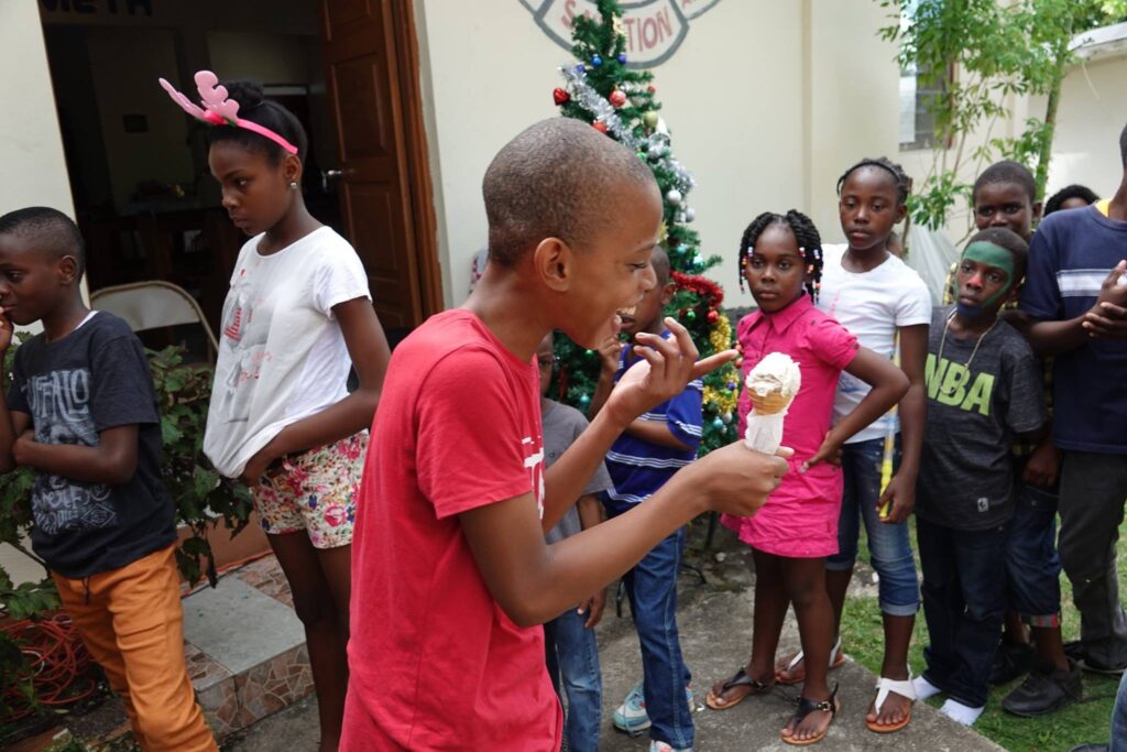 Children gather around a boy enjoying ice cream, smiling and animated, near a Christmas tree, highlighting a festive celebration.