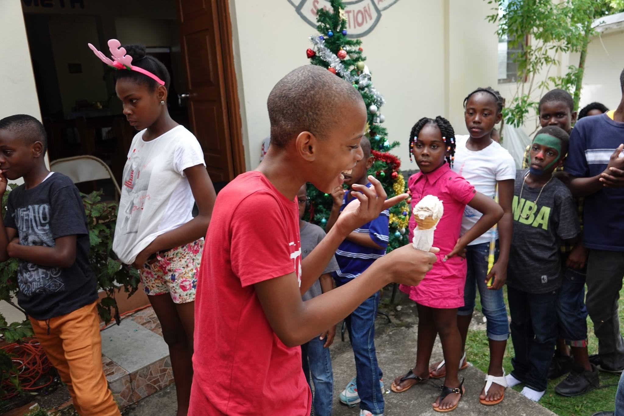 Children gather around a boy enjoying ice cream, smiling and animated, near a Christmas tree, highlighting a festive celebration.
