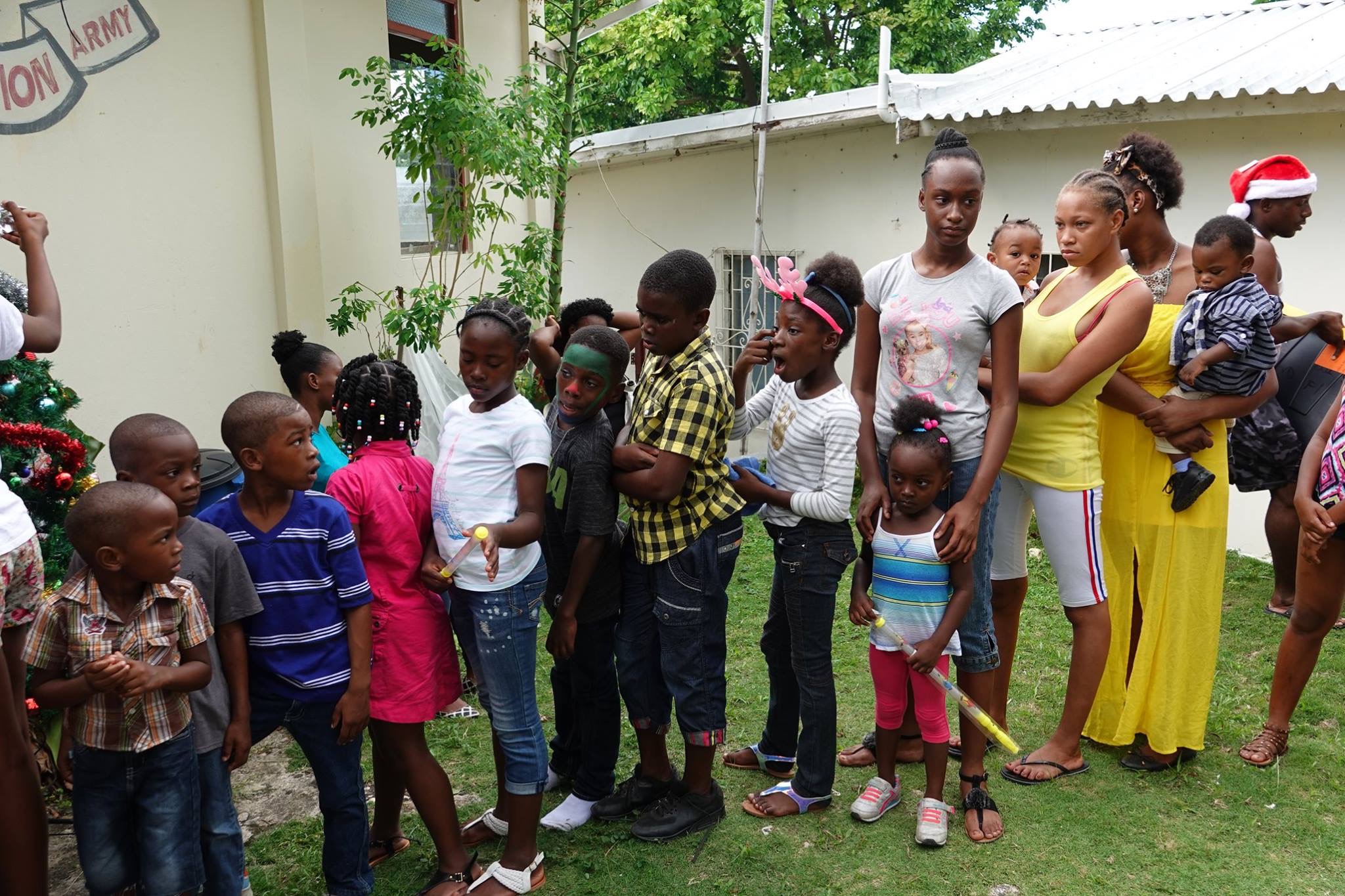 Children and adults gather for a festive event outside, with a Christmas tree nearby. Joyful expressions and colorful outfits enhance the lively atmosphere.