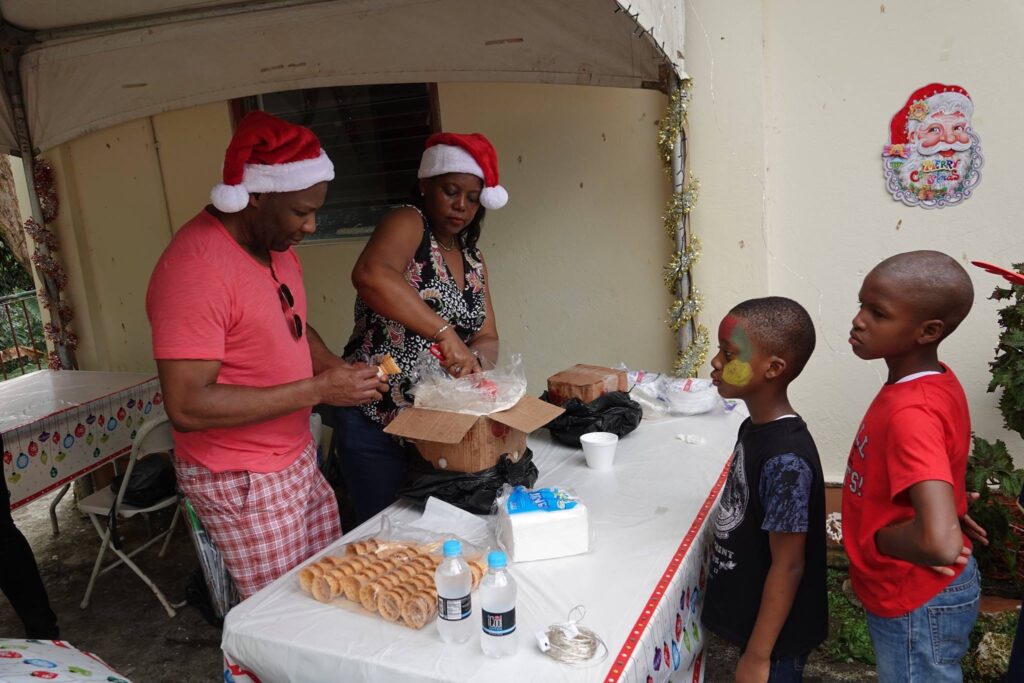 A festive scene featuring a man and woman in Santa hats preparing food at a table, with two children observing. The atmosphere is celebratory, hinting at a holiday gathering.