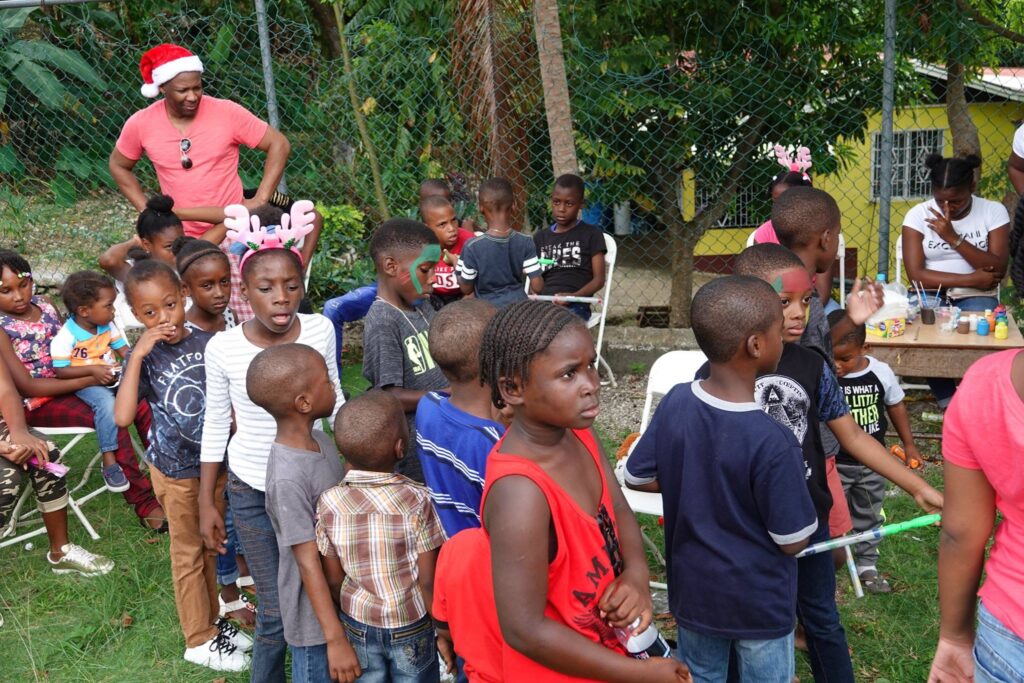 Children gather at a festive outdoor event, with some wearing holiday-themed accessories. The scene captures their excitement and community spirit.