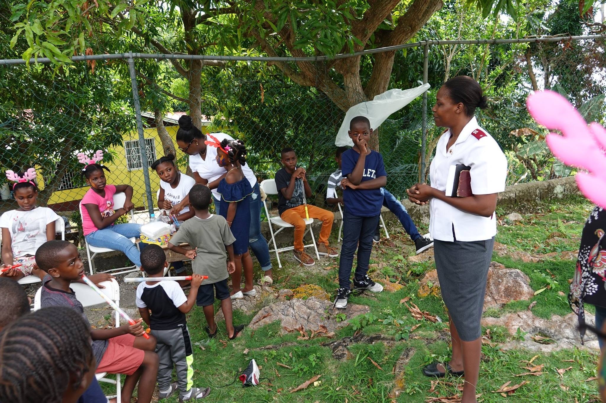 Children and adults gather outdoors for a community event, with a woman in a uniform speaking while others engage in activities, promoting connection and support.