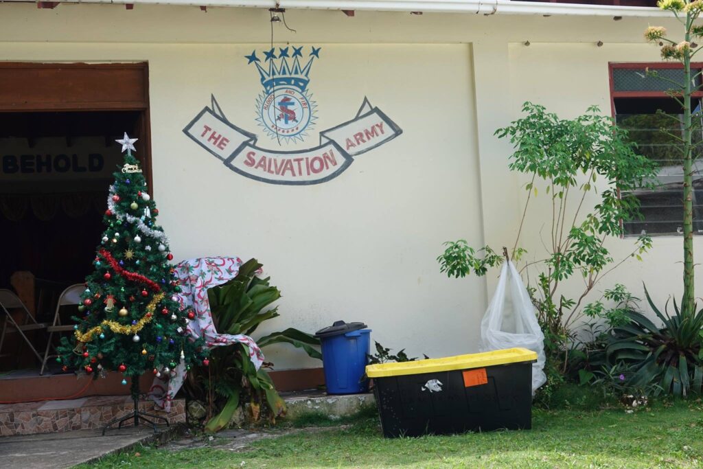 Christmas tree adorned with decorations stands outside a Salvation Army building, featuring a large logo on the wall and surrounding greenery.