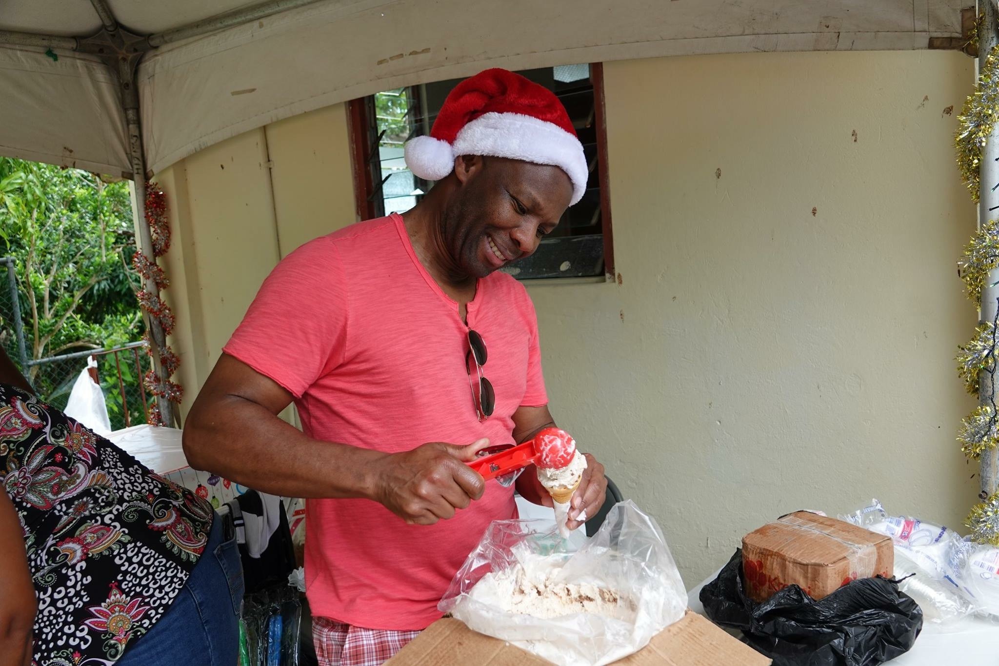 A man in a Santa hat joyfully prepares ice cream cones at a festive outdoor event, highlighting a communal celebration of holiday spirit.