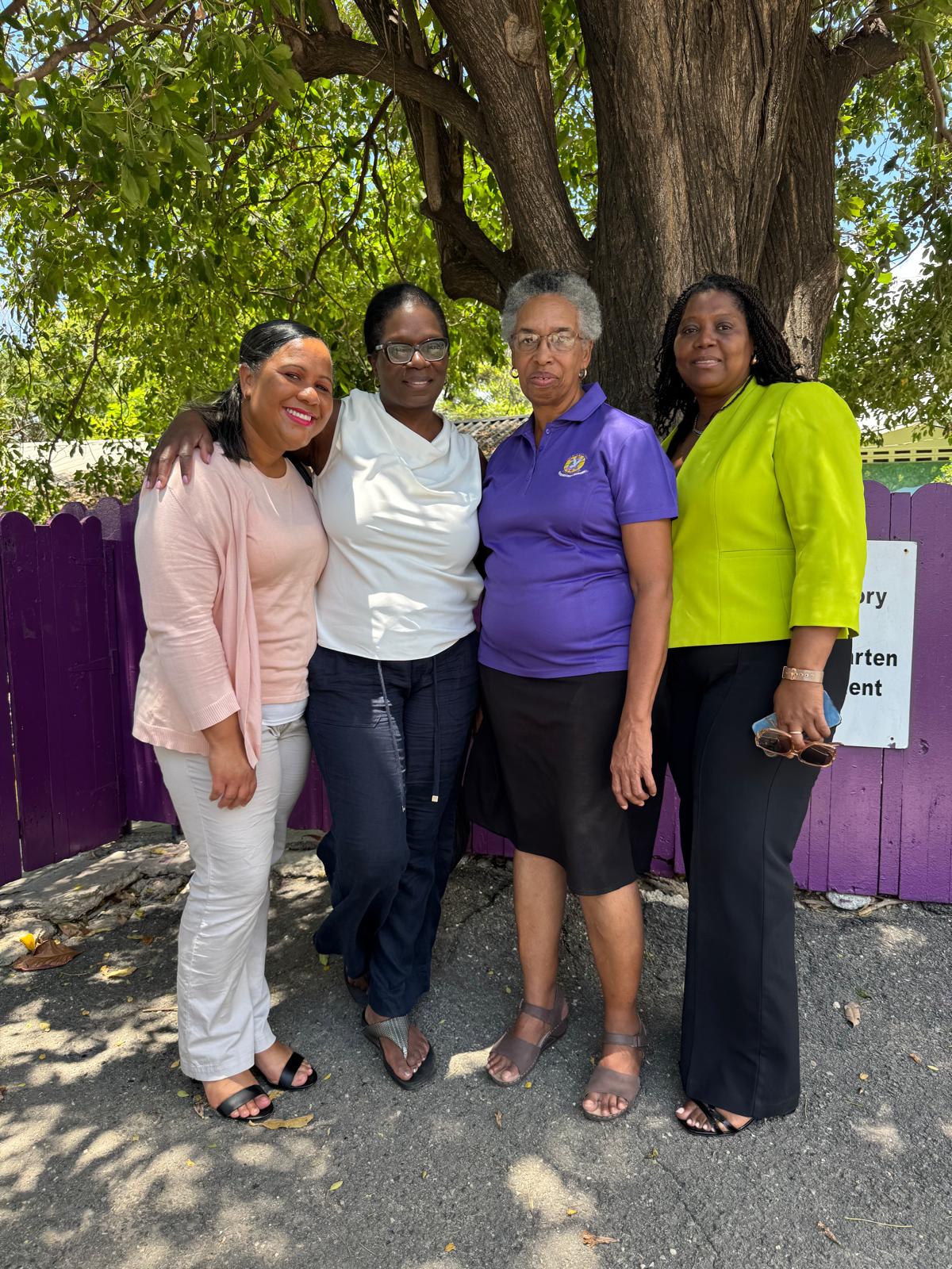 Four women stand together under a leafy tree, smiling and dressed in colorful outfits. The scene conveys unity and camaraderie.