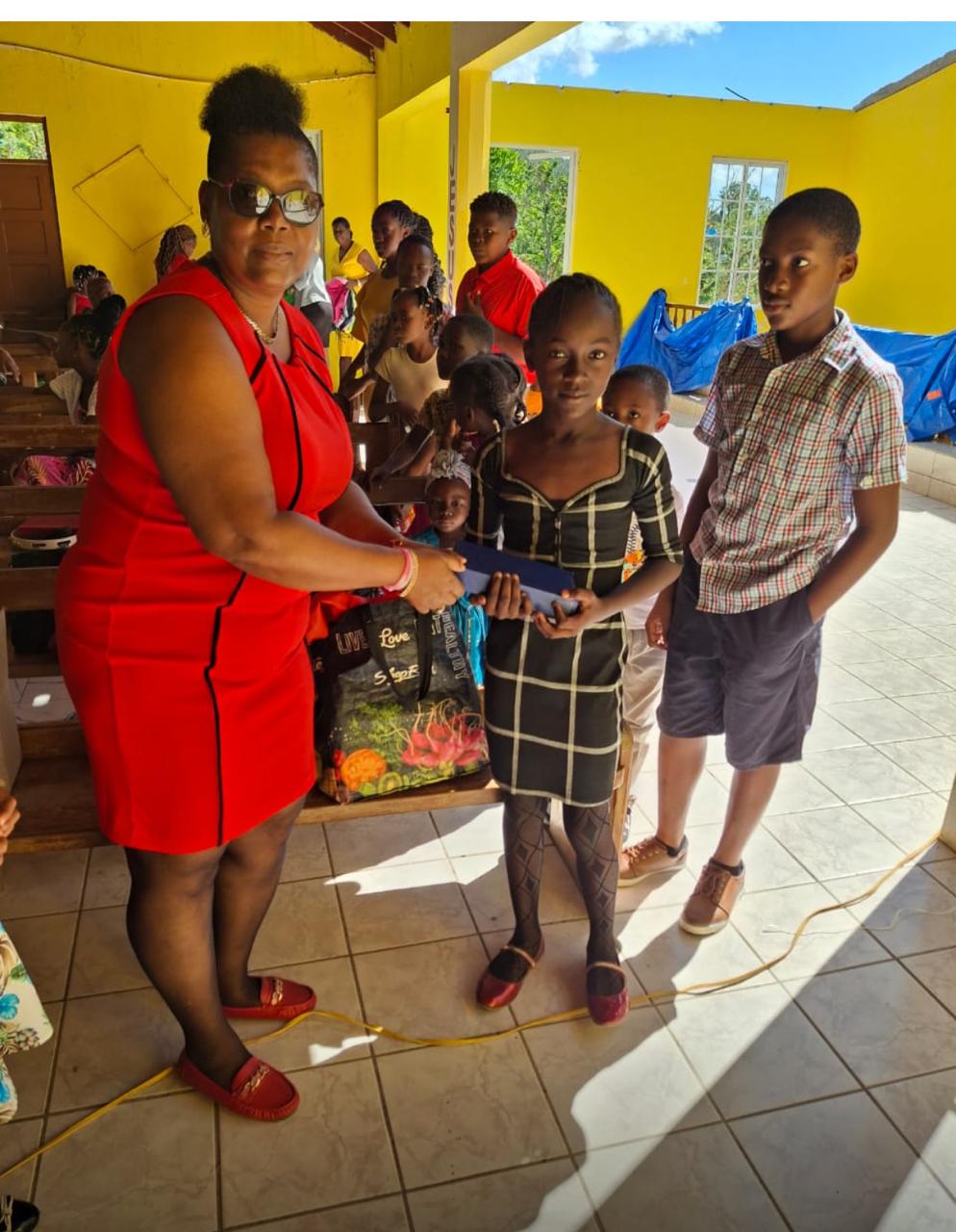 A woman in a red dress presents a blue item to a young girl in a plaid dress, while other children observe in a brightly lit community space.