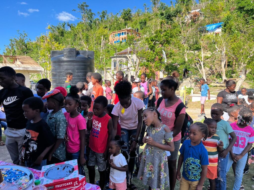Children gather joyfully at an outdoor event, awaiting treats at tables adorned with festive snacks like fruitcake, surrounded by greenery and bright skies.