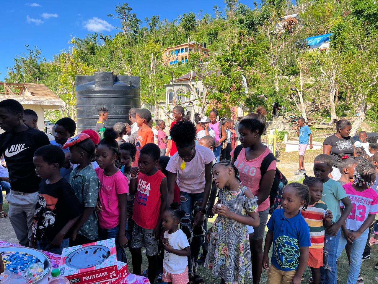 Children gather joyfully at an outdoor event, awaiting treats at tables adorned with festive snacks like fruitcake, surrounded by greenery and bright skies.